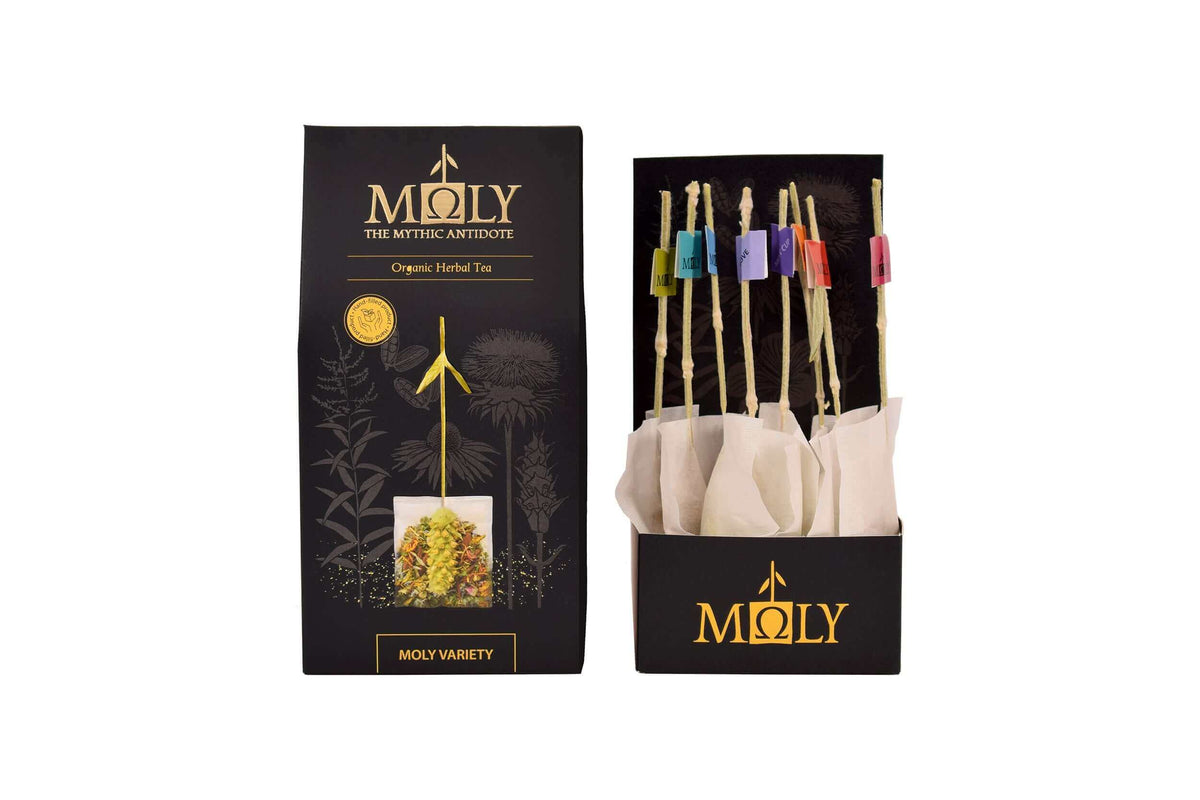 Eine schwarze Verpackung des MOLY VARIETY BIO sowie aufgedruckten Pflanzen. Zentral ist Bild aufgedruckt, welches einen Teebeutel mit Inhalt zeigt aus dem der Stengel der Sideritis scardia (griechischer Bergtee) herausragt. Daneben ist der Inhalt der Verpackung zu sehen, der aus zehn einzelnen Teebeuteln mit jeweils einem Sideritis-Stengel besteht.