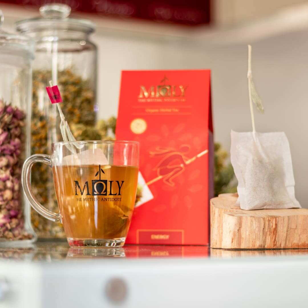 Eine rote Verpackung des Moly Energy Bio vor einem Hintergrund mit Vorratsgläsern an Kräutern. Zentral ist ein Bild aufgedruckt, welches einen Teebeutel mit Inhalt zeigt aus dem der Stengel der Sideritis scardia (griechischer Bergtee) herausragt und auf dem eine gezeichnete Hexe fliegt. Auf der linken Seite steht ein Tee aus dem der Sideritis -Stengel herausragt. Rechts ist der Teebeutel zu sehen.