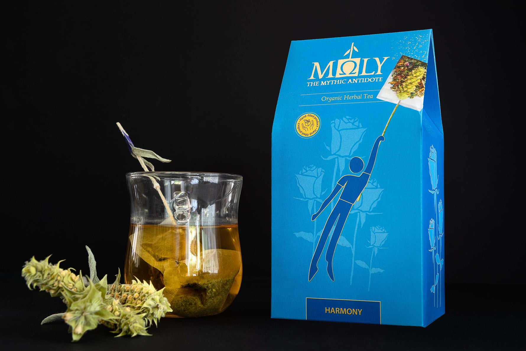 Eine blaue Verpackung des Moly Harmony BIO vor einem schwarzen Hintergrund. Zentral ist ein Bild aufgedruckt, welches einen Teebeutel mit Inhalt zeigt, aus dem der Stengel der Sideritis scardia (griechischer Bergtee) herausragt und auf dem eine Person vom Tee in die Luft gezogen wird. Auf der linken Seite steht ein Tee aus dem der Sideritis -Stengel herausragt. Davor liegen zwei Sideritis-Blüten.